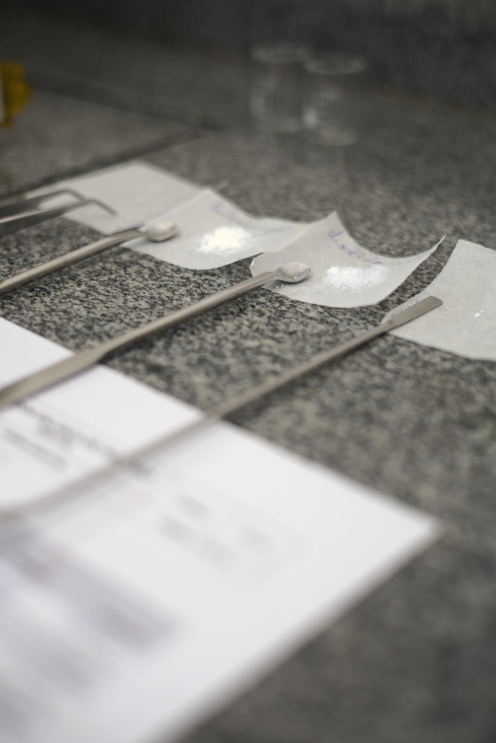 Imagem de três amostras de matéria-prima em pó, dispostas em pequenas espátulas sobre papéis. A imagem representa o estudo de equivalência para controle de qualidade e segurança.